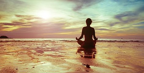 Foto de uma senhora a meditar em frente ao mar calmo e com um ceu ao fundo cor de laranja. 